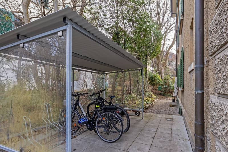 A covered outdoor area with a metal roof and glass sides, featuring several bicycles parked inside. The area is adjacent to a building with a stone wall and a green door, surrounded by trees and greenery.