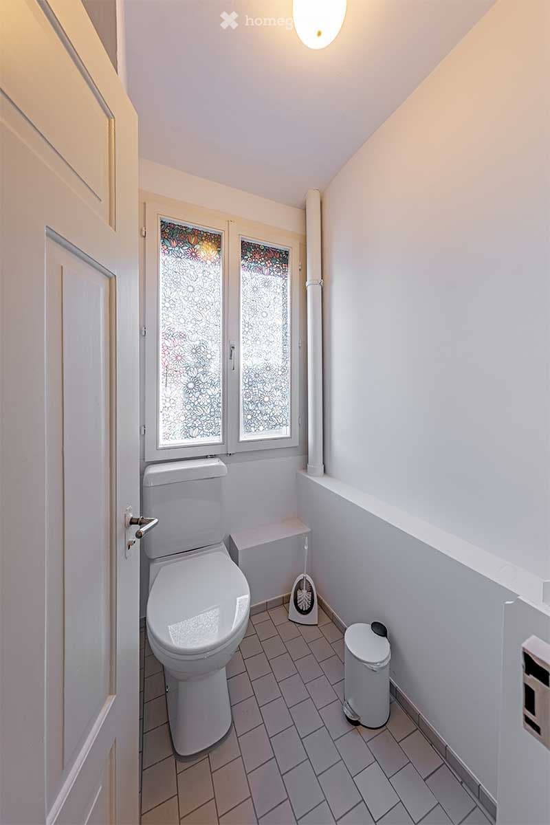 A bathroom with a toilet, white tiled floor, and white walls. There are two windows with glass panes and a door leading to the outside.