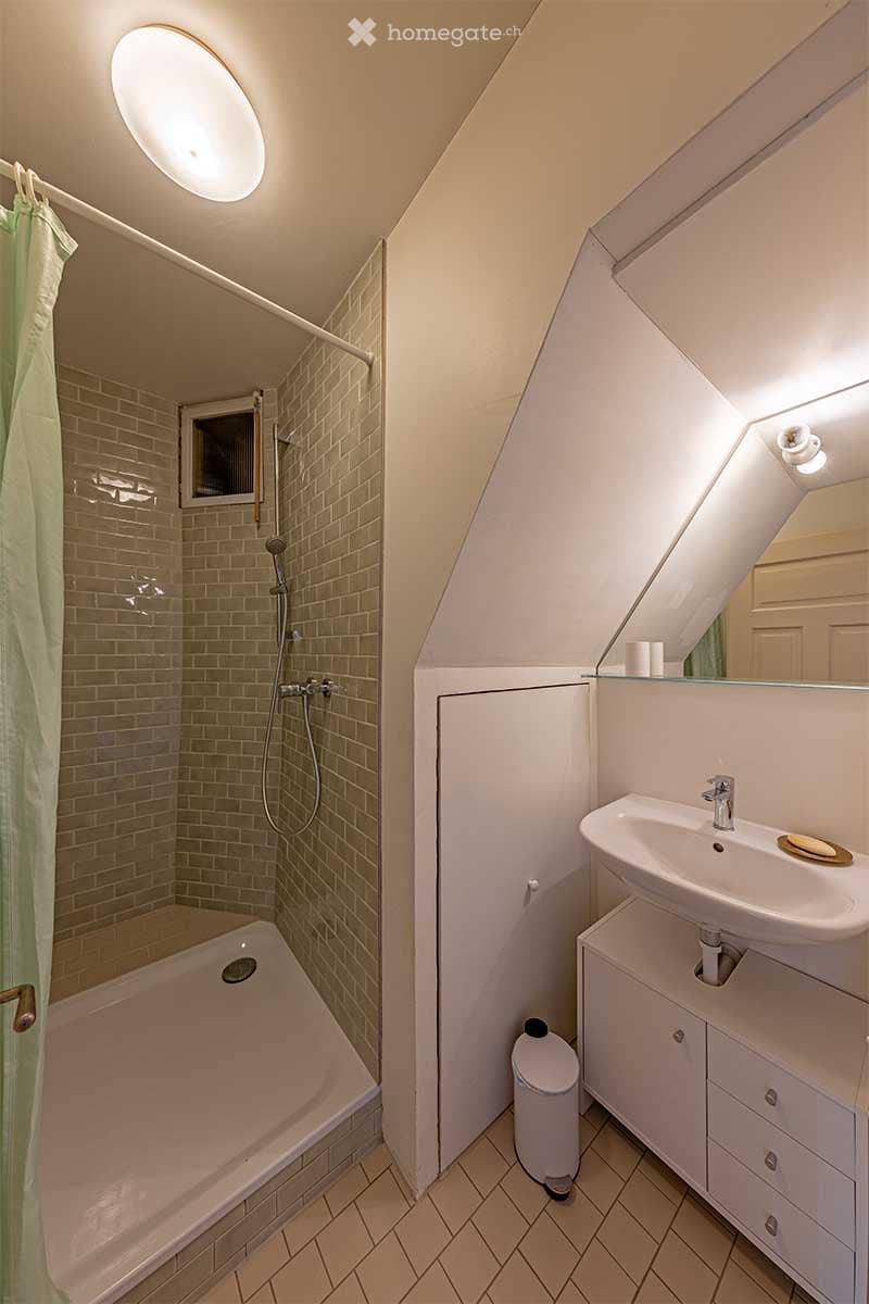 The bathroom has a shower with glass doors, a bathtub, a sink with a faucet, a mirror, white tiles, and a waste bin. It also has a small window for ventilation.