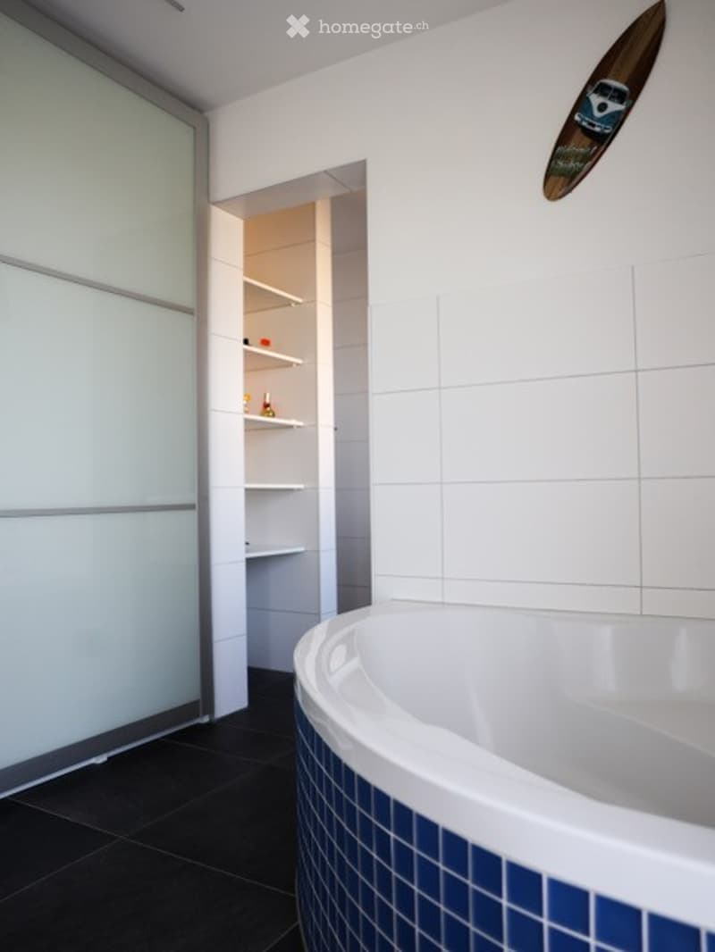 Modern bathroom with a white bathtub featuring blue tiles, dark tiled floor, and an open shelf unit with various items.