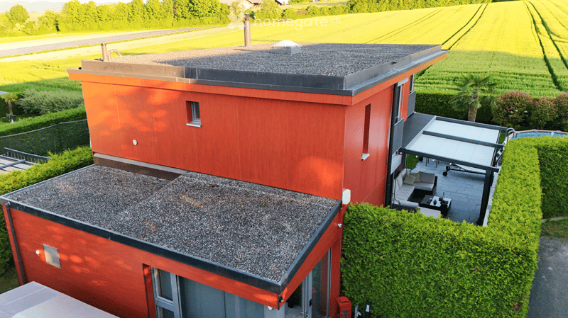 Modern two-story house with red exterior and grey gravel roofs, featuring a patio with outdoor furniture and a swimming pool.