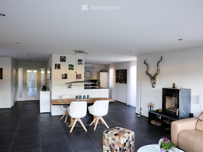 Modern living room with a beige sofa, colorful patterned stool, dining table with four chairs, fireplace, and artwork on the walls.