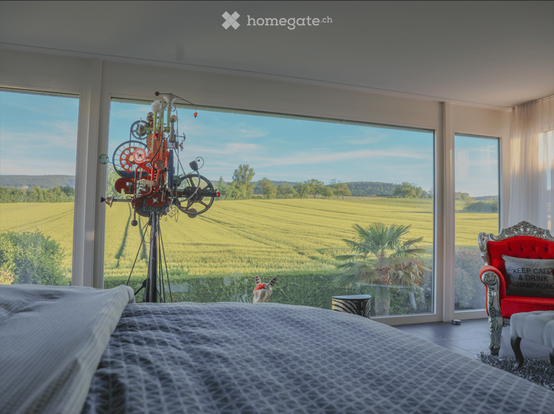 Bright bedroom with large windows overlooking a scenic countryside view, featuring a bed, a red armchair, a small table, and a colorful kinetic sculpture.