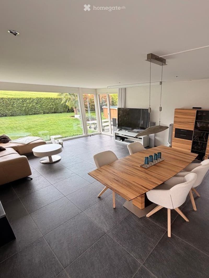 Open-plan living and dining area with large windows offering a view of a green garden. Features a grey sofa, coffee table, wooden dining table with white chairs, and a TV on a stand.