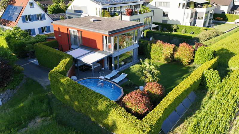 Aerial view of a modern house with a swimming pool, surrounded by well-maintained hedges and greenery, with neighboring houses visible in the background.