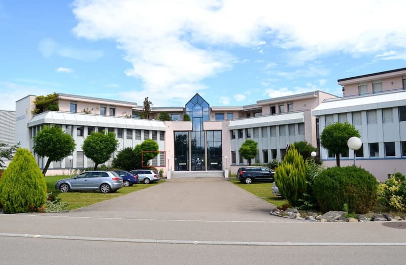 A modern, multi-story office building with a glass atrium entrance. The building has a symmetrical design with wings on either side. There are several cars parked in the front of the building, indicating a parking area. The building is surrounded by lands