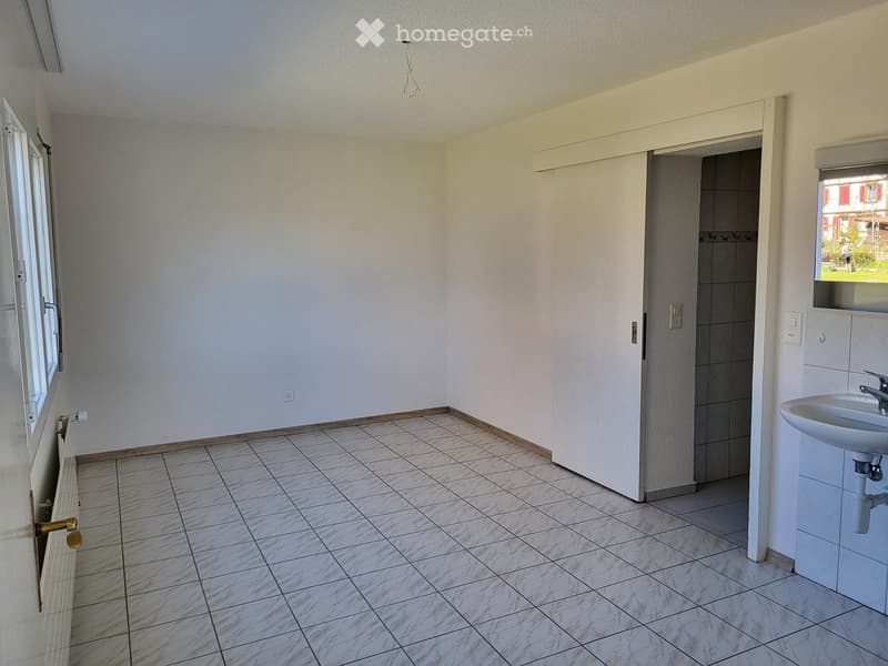 Empty room with tiled floor, white walls, and a small sink in the corner. There is a door leading to another room and a window with a view of the outside.