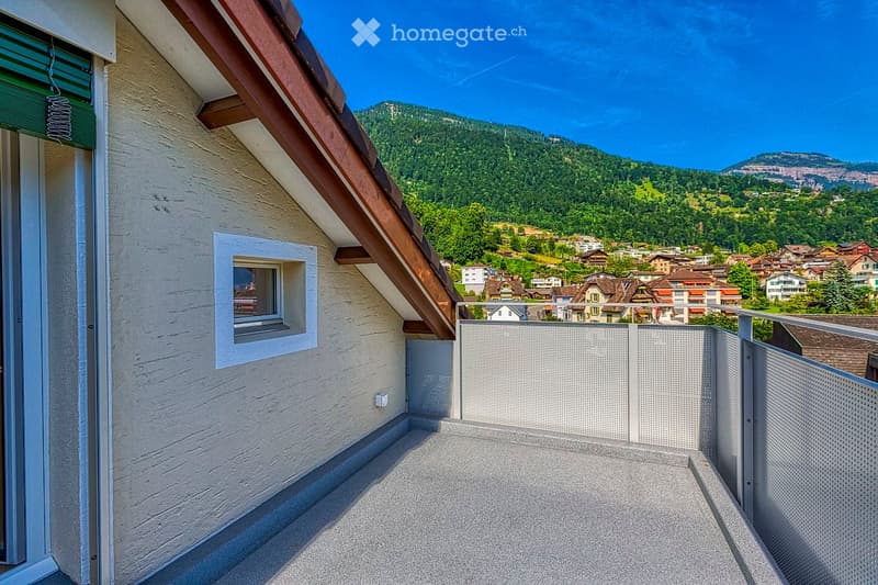 Balcony with a view of a town and mountains, featuring a metal railing and a small window on the adjacent wall.