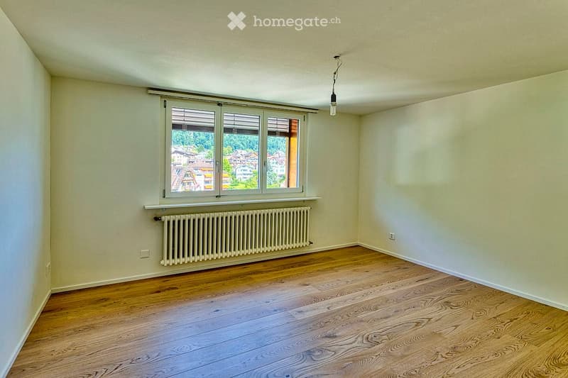 Empty room with wooden flooring, white walls, large window with blinds, radiator, and a single hanging light fixture.