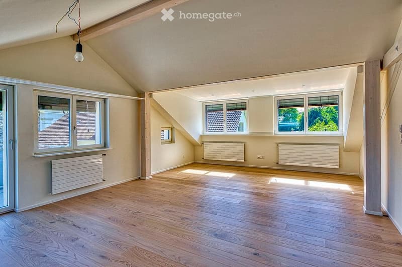 Bright, empty attic room with wooden flooring, large windows, radiators, and exposed wooden beams on the ceiling.