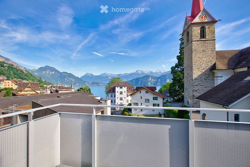 Balcony overlooking a town with a church tower, houses, and a lake surrounded by mountains.