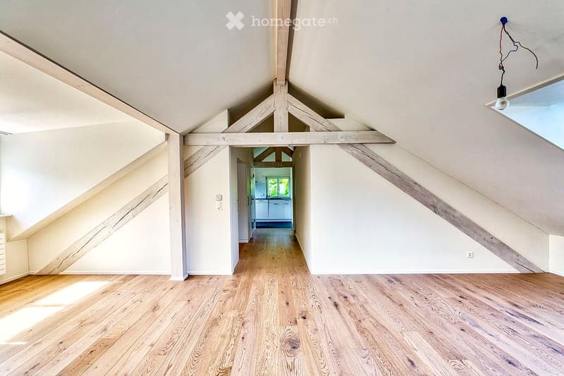 An empty attic space with wooden floors, exposed wooden beams, and natural light coming through a skylight. The room has white walls and a doorway leading to another room.