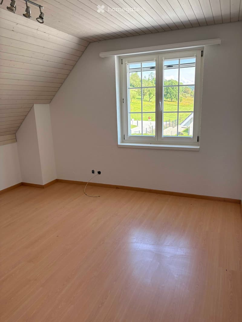 Empty room with wooden flooring, white walls, and a large window offering a view of a green landscape.