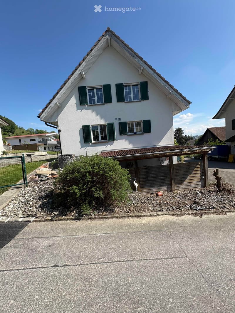 Two-story house with white exterior, green shutters, and a pitched roof. The house features multiple windows and a small covered entrance area with a wooden fence and decorative rocks.