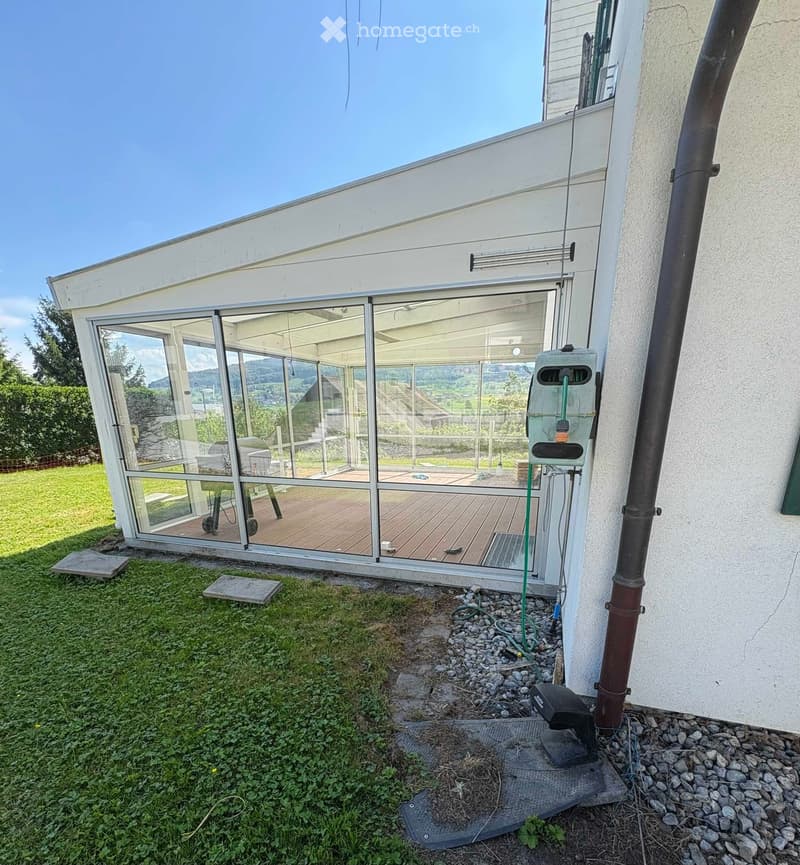 A modern glass-enclosed patio attached to a house, with wooden flooring, a view of the surrounding landscape, and some outdoor equipment visible near the entrance.