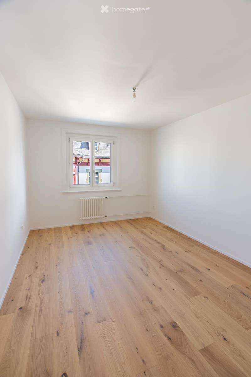 Empty room with light wooden flooring, white walls, a single window, and a radiator below it.