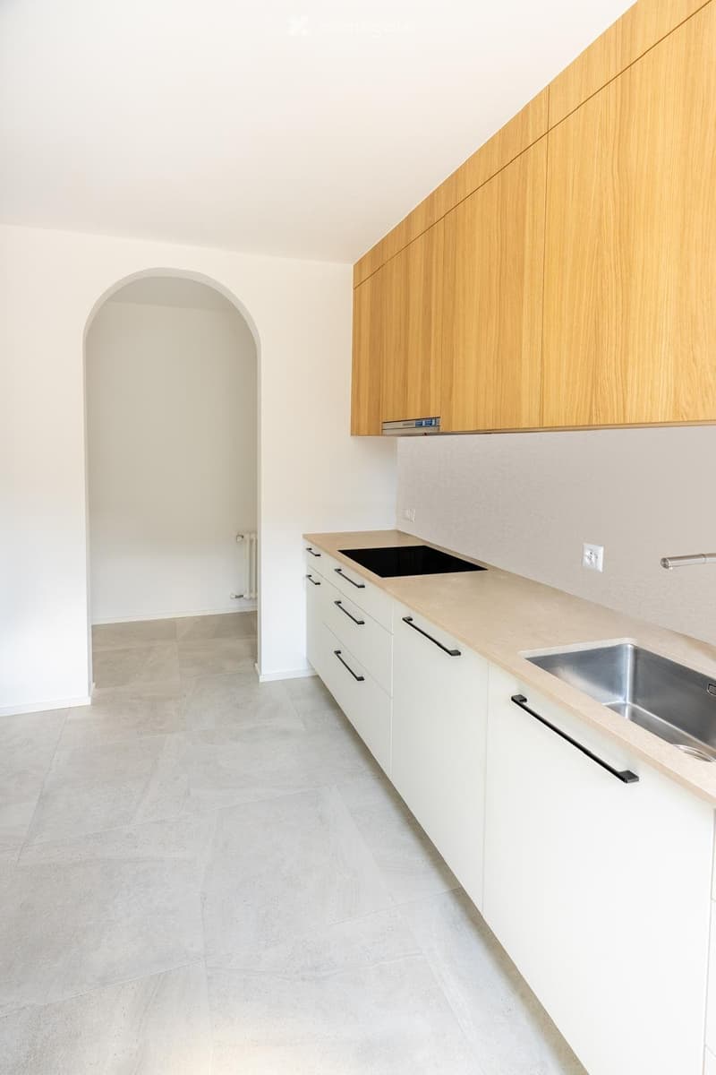 Modern kitchen with light wooden cabinets, white countertops, a sink, and a tiled floor. The space is clean and well-lit with an arched doorway leading to another room.