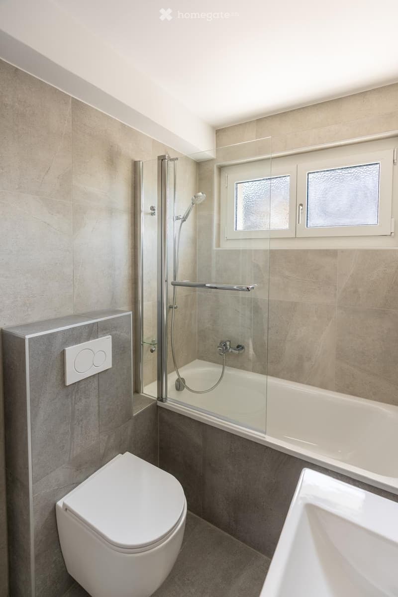 Modern bathroom with a toilet, bathtub with glass shower enclosure, and a sink. The walls are tiled in a neutral color, and there is a window with frosted glass above the bathtub.