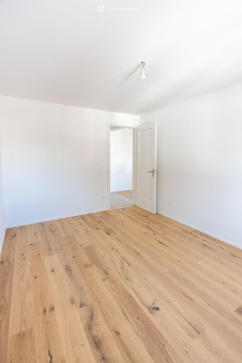 Empty room with light wooden flooring and white walls, featuring a single open door leading to another space.
