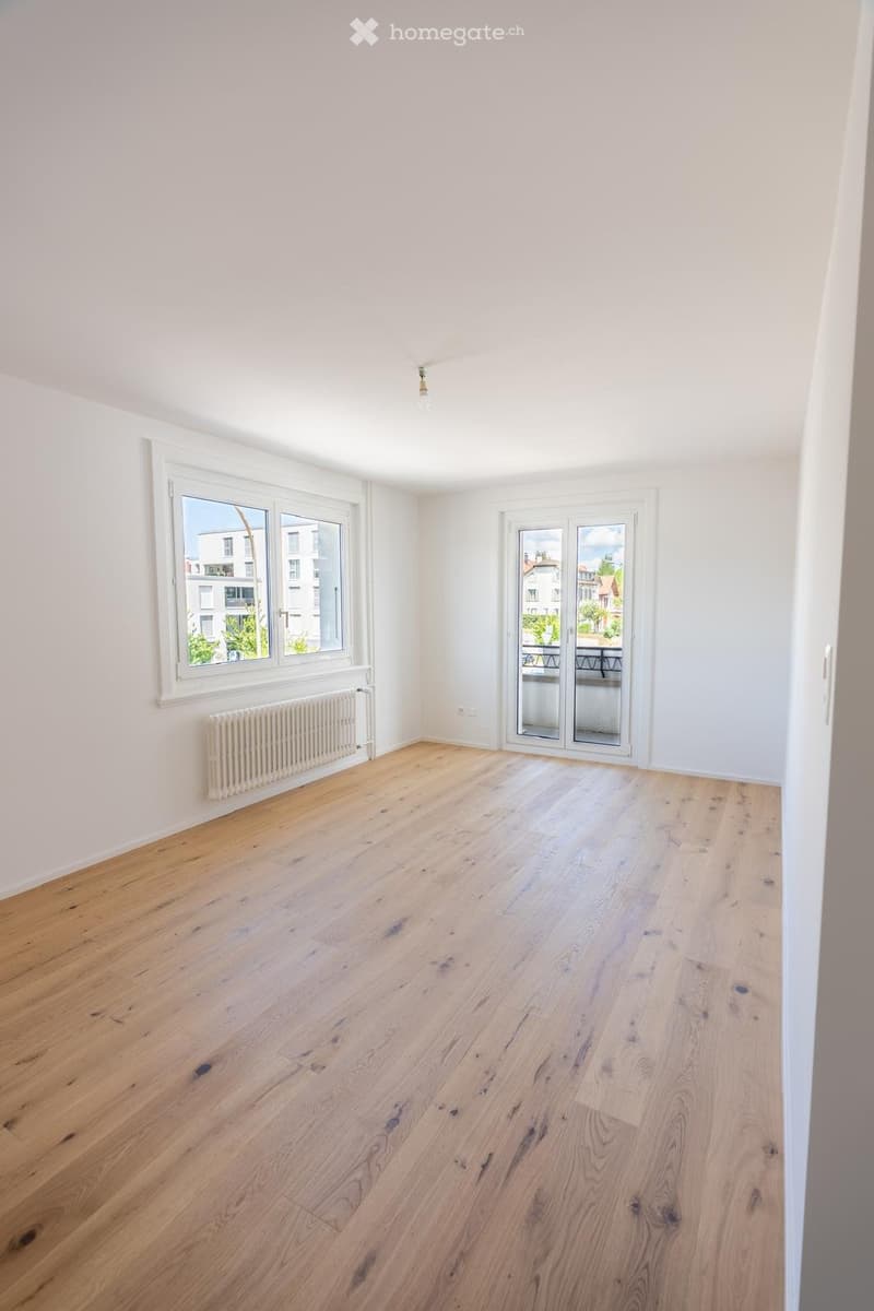 Empty room with light wooden flooring, white walls, large windows, and a sliding glass door leading to a balcony.