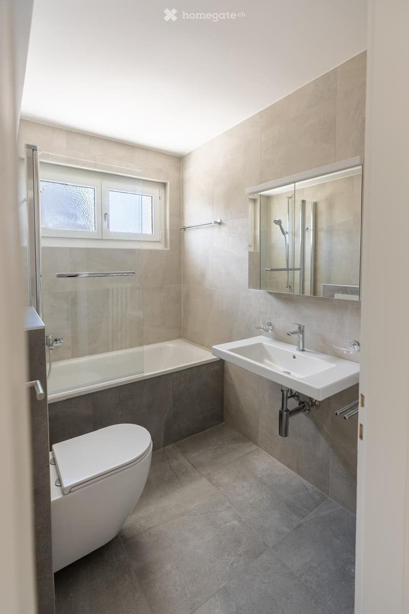 Modern bathroom with a toilet, sink, bathtub, and large mirror. Features tiled walls and floor, a window, and modern fixtures.