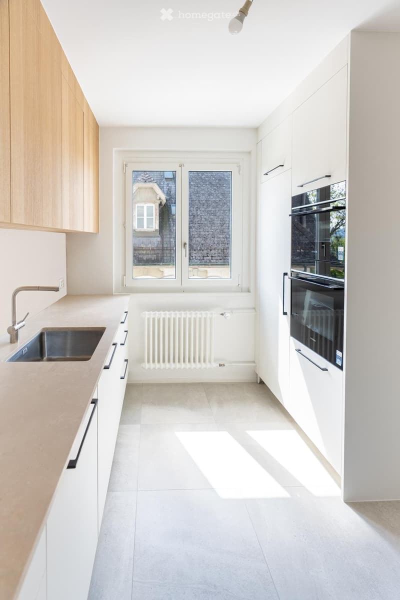 Modern, minimalist kitchen with white cabinets, a stainless steel sink, a built-in oven, and a radiator. The space features large windows and a clean, uncluttered design.