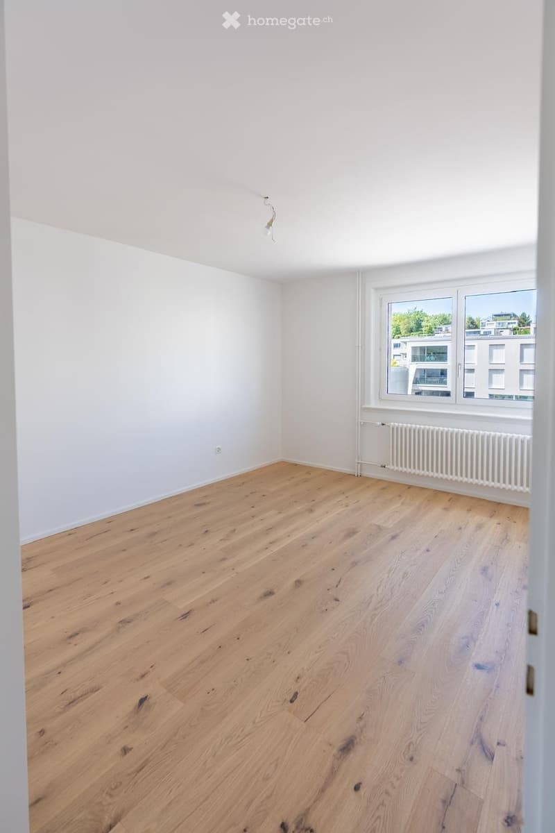 Empty room with light wooden flooring, white walls, and a large window with a radiator below it.
