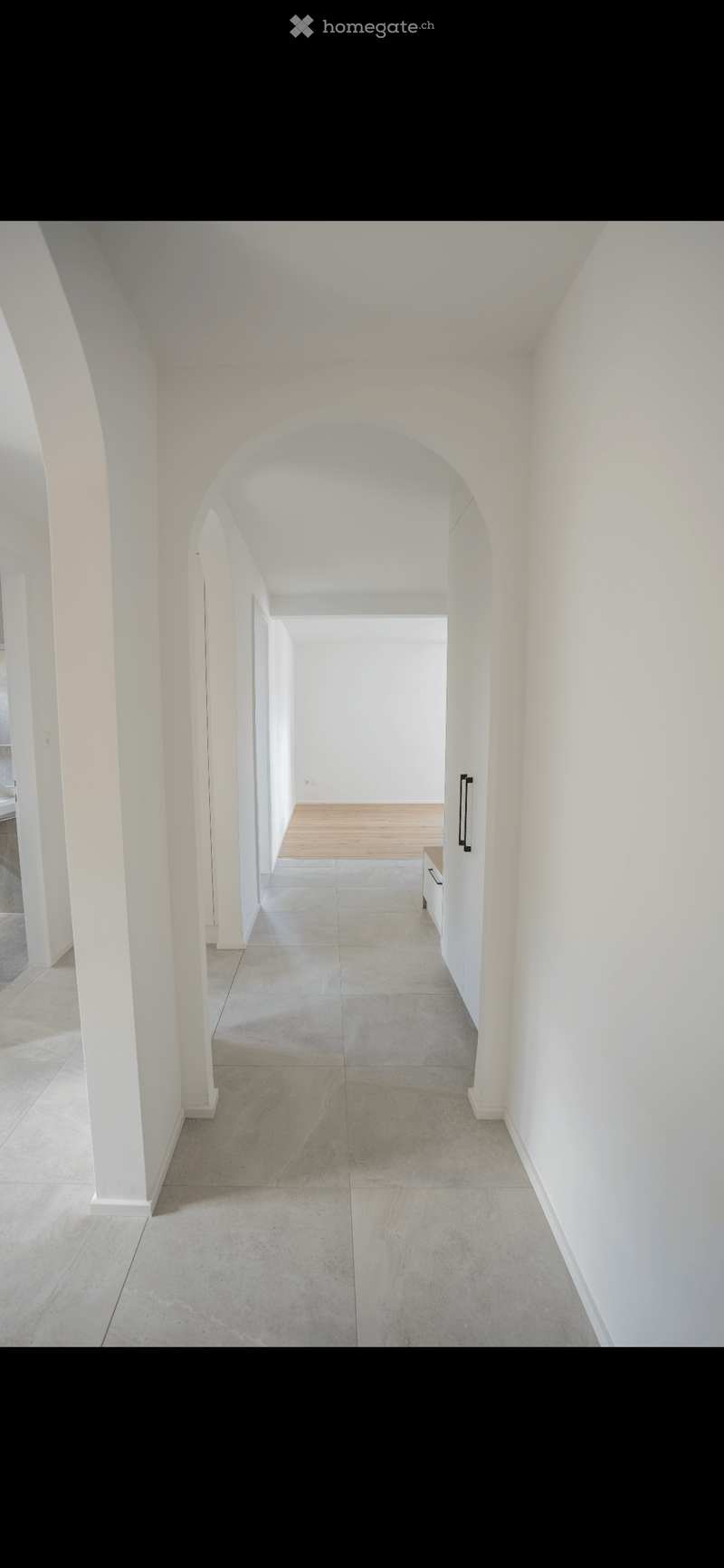A long, white-walled corridor with arched doorways, polished concrete floors, and minimal furnishings.