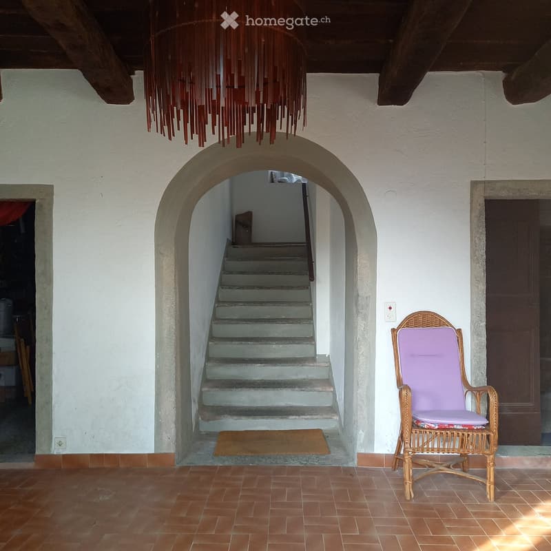 A spacious, well-lit hallway with a large arched doorway leading to a staircase. The floor is tiled in warm terracotta, and a single wicker chair with a purple cushion sits near the doorway.