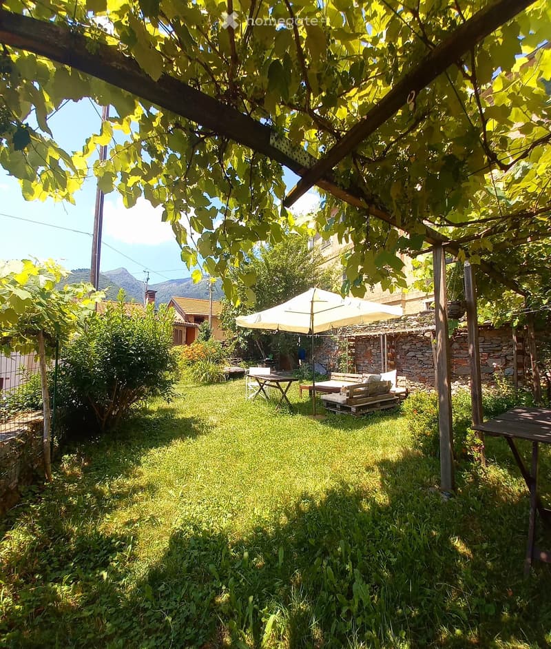 A lush garden with a wooden pergola, outdoor seating, and a stone wall, surrounded by greenery and a scenic mountain view.