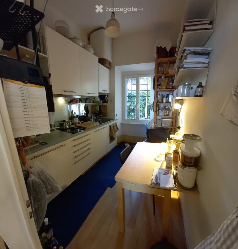 A compact kitchen with white cabinets, a green countertop, and a wooden island. Visible appliances include a refrigerator, microwave, oven, and blender. Shelves hold various kitchen items and books.