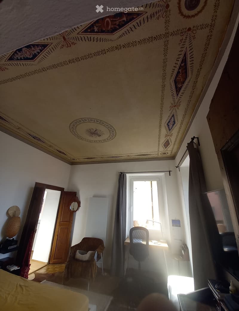 Bedroom with a bed, armchair, desk, chair, mirror, and a window with curtains. The room has ornate ceiling decorations and a door leading to another space.