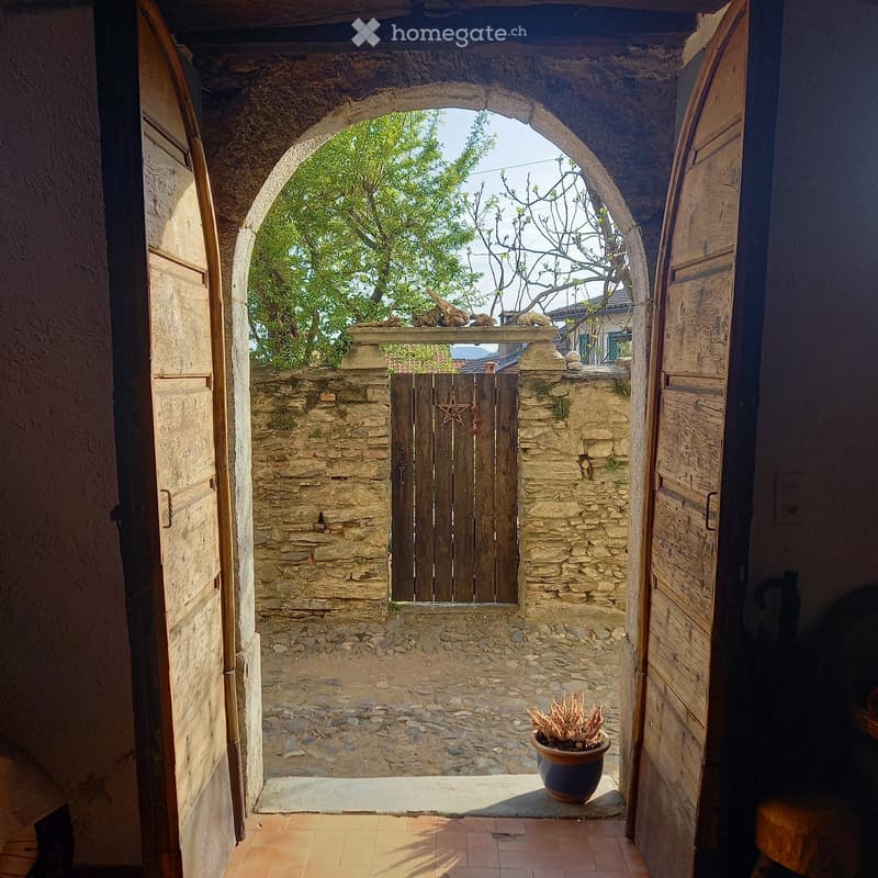 An arched stone doorway leads to an outdoor courtyard with a stone wall, wooden gate, and potted plant.