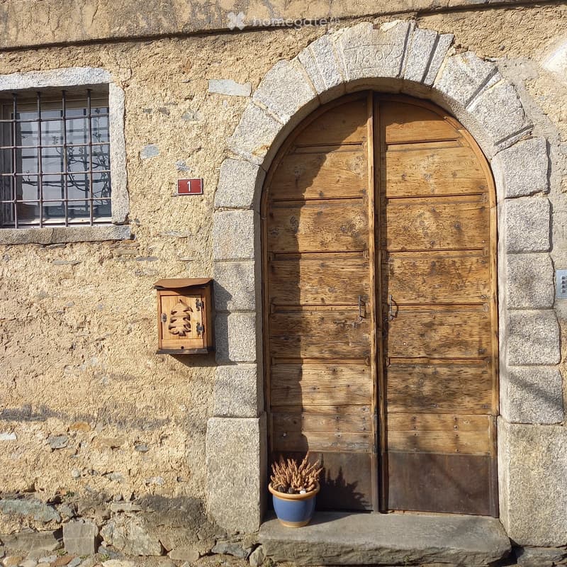Stone building entrance with a wooden double door, a small window with metal bars, a mailbox, and a potted plant on the doorstep.