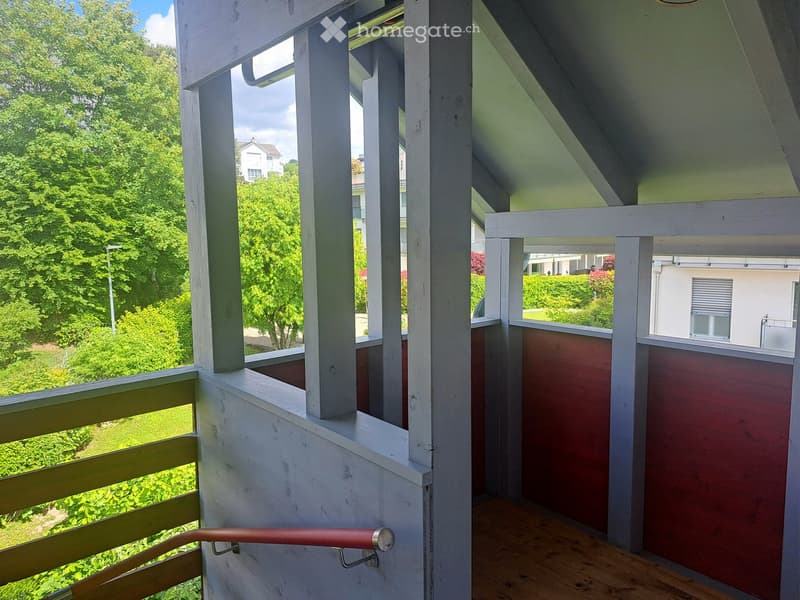 Wooden balcony with white railings, wooden floor, and red handrail, overlooking a green garden and neighboring houses.