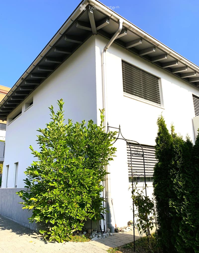 White two-story house with modern design, featuring multiple windows with blinds, a covered entrance area, and surrounding greenery including a bush and a climbing plant on a metal trellis.