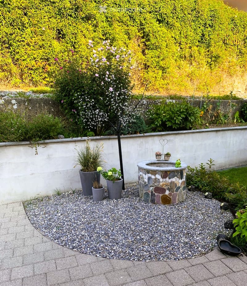 A small garden area with a stone fountain, potted plants, and a gravel bed surrounded by a low wall and lush greenery.