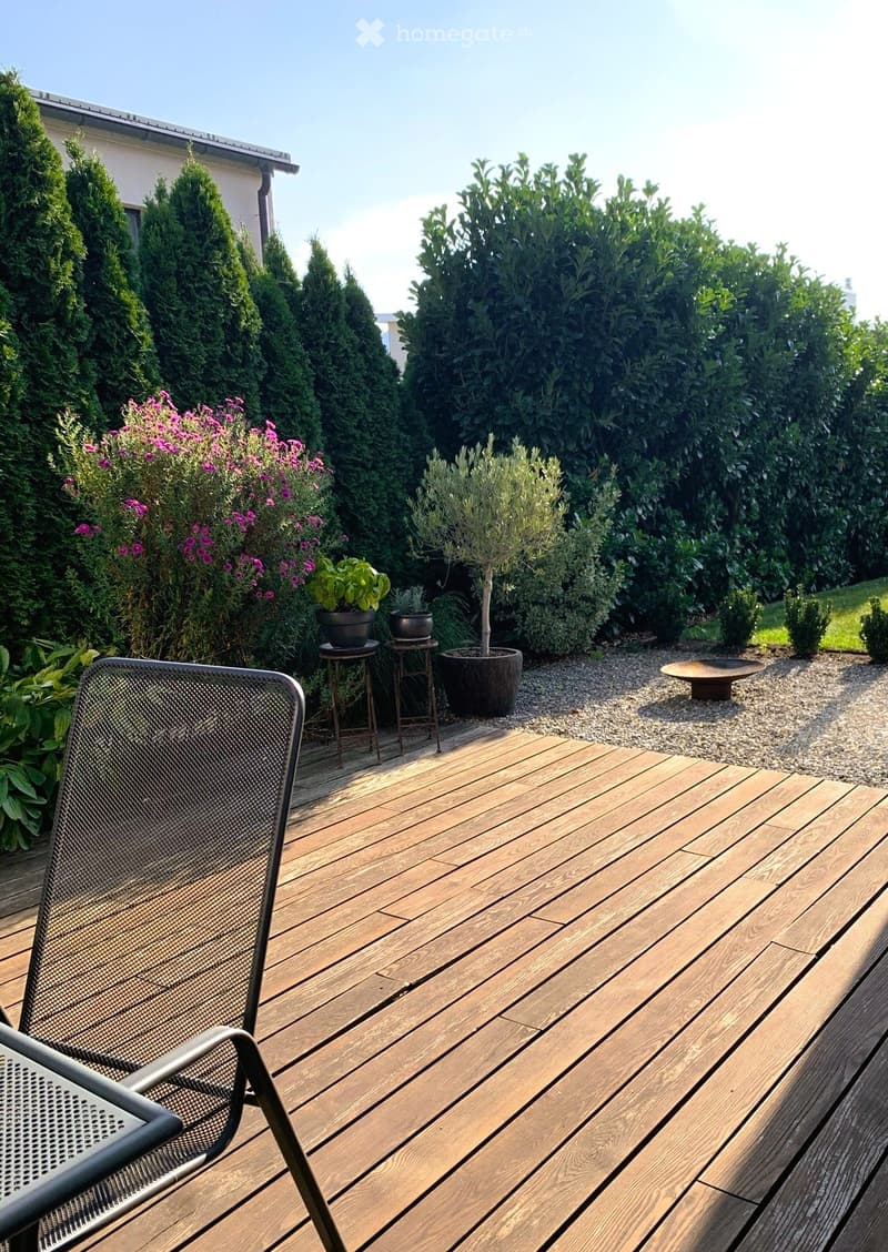 Wooden deck with a metal chair, surrounded by potted plants, shrubs, and a hedge. The deck is bordered by gravel and features a small round table and additional potted plants.