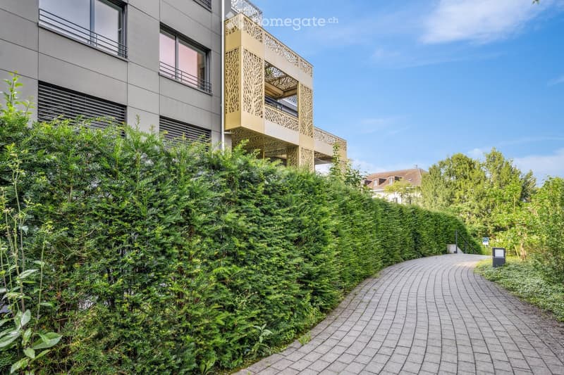 Garden / Outdoor Area. A modern building with a decorative balcony is partially visible behind a lush green hedge. A paved pathway winds through the garden area, leading towards the building.