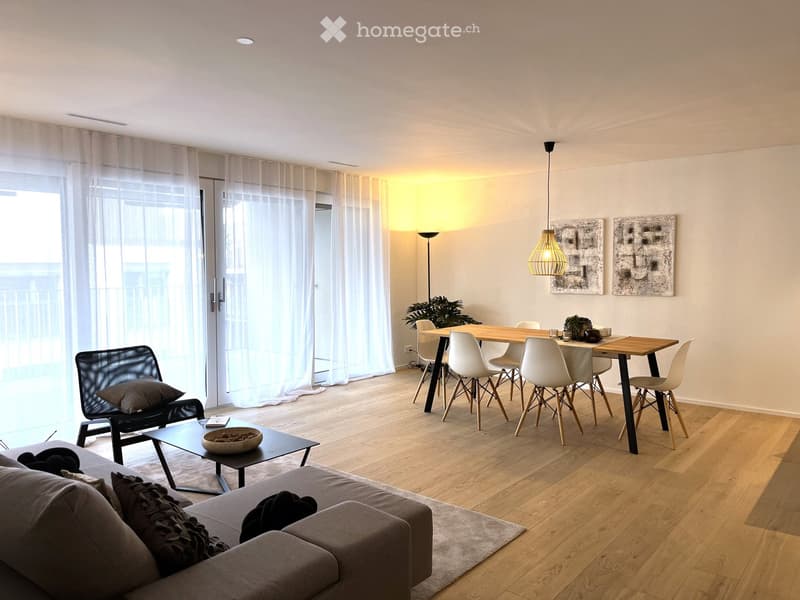 Open floor plan with a dining area, a living area with a couch and armchair, white walls, wooden floor, glass doors leading to a balcony, and white curtains