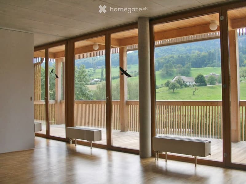 Bright, open-plan room with large sliding glass doors leading to a wooden deck, offering scenic countryside views. The room features a polished wooden floor and two radiators placed near the glass doors.