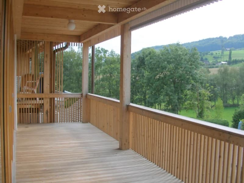 Wooden balcony with open railings, offering a scenic view of a green landscape and hills.