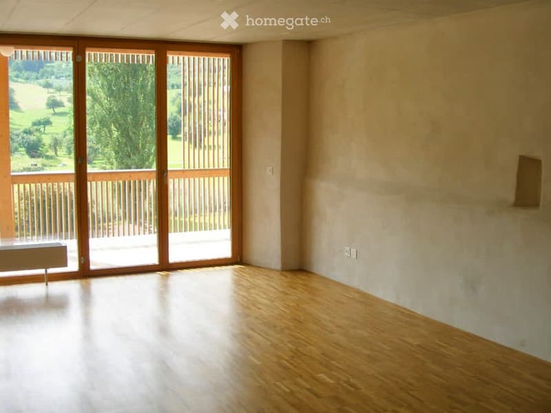 Empty room with wooden flooring, large sliding glass doors leading to a balcony with a scenic view of greenery and a field.