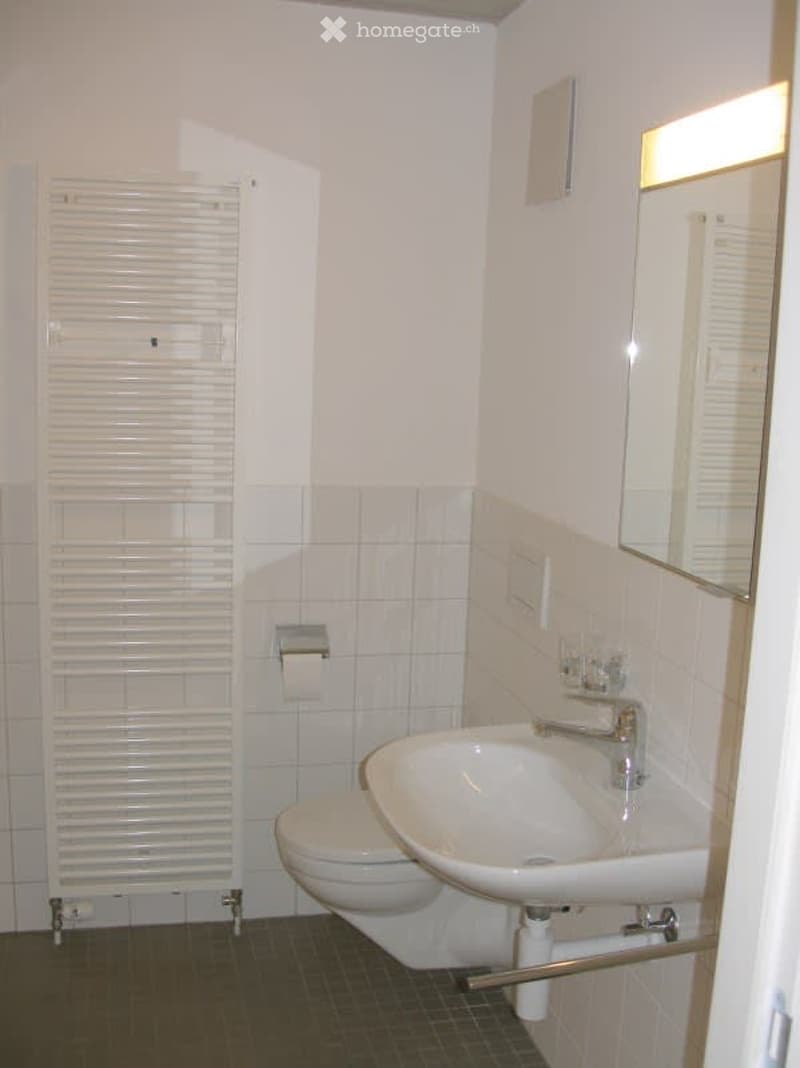 A small, clean bathroom with white tiled walls, a toilet, a sink with a mirror above it, and a towel rack on the left side.