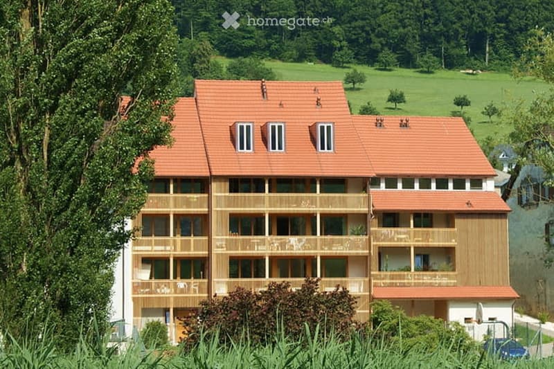 Multi-story residential building with red-tiled roof, multiple balconies, and large windows, surrounded by greenery and trees.