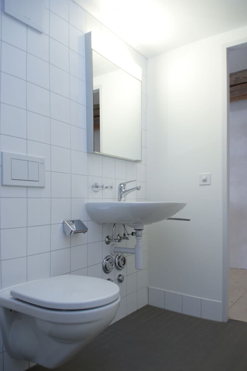 Modern bathroom with white tiled walls, a toilet, a sink with a chrome faucet, and a large mirror above the sink. The floor is dark tiled.