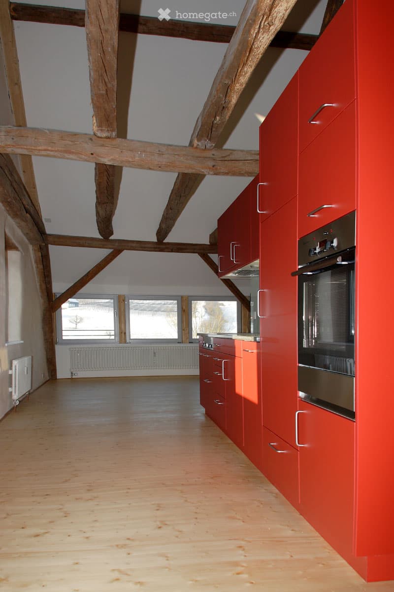 Bright kitchen with exposed wooden beams, red cabinets, and a stainless steel oven. The floor is light wooden, and there are large windows letting in natural light.
