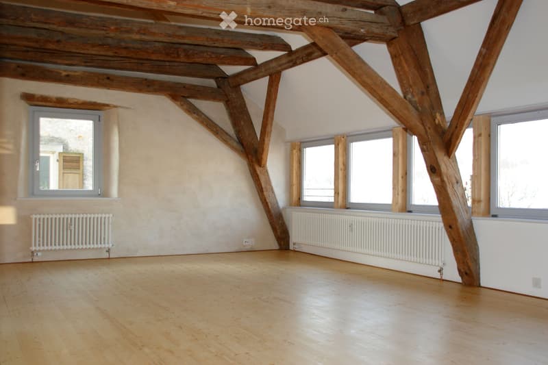 Empty attic room with wooden beams, wooden floor, and large windows. No furniture or equipment.