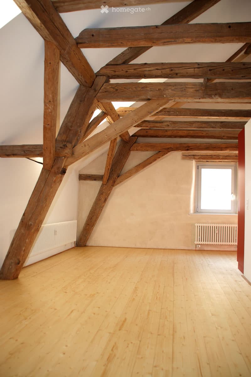 Empty attic room with exposed wooden beams, wooden flooring, and a single window letting in natural light.