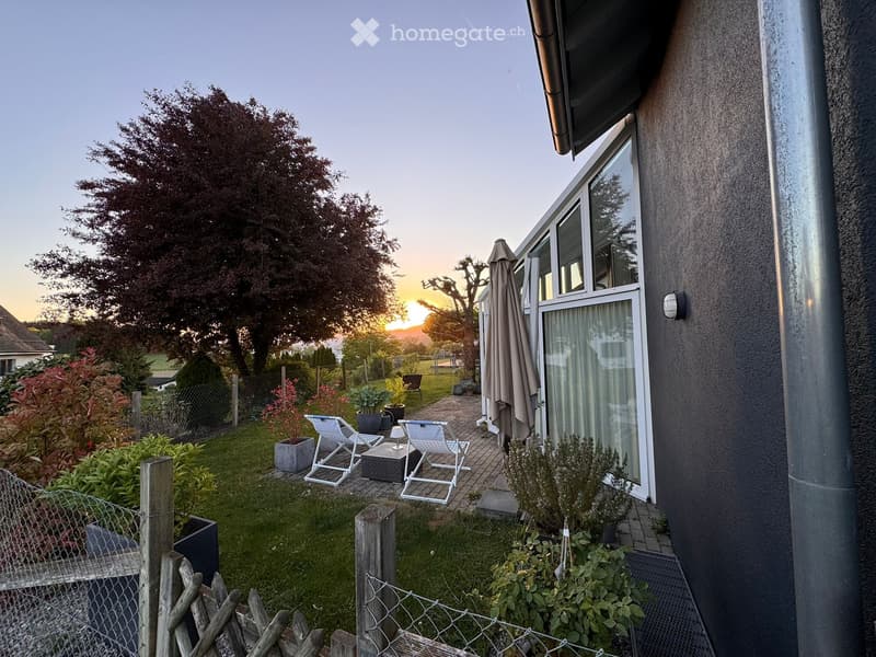 Outdoor area with a patio, chairs, a table, and a closed umbrella. The patio is surrounded by a fence and has a grassy lawn. There are various plants and trees in the area, and a building is visible in the background. The sky is clear with a sunset in the